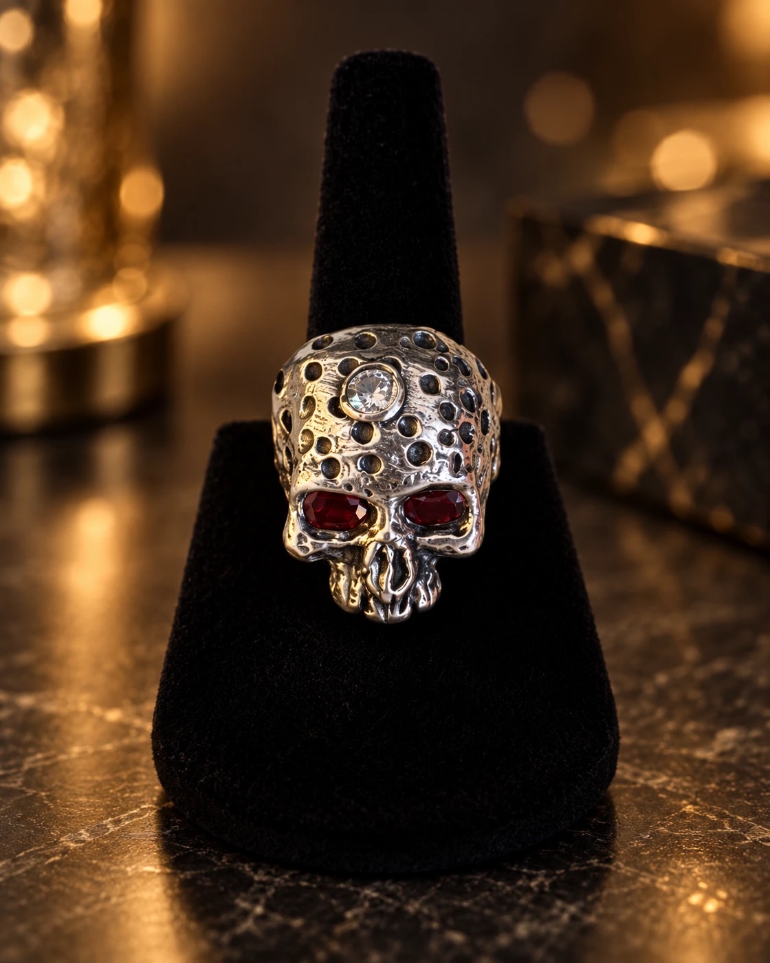 Hand-carved silver skull ring with pitted texture, red stone eyes and a clear stone set in the crown, on a black velvet stand